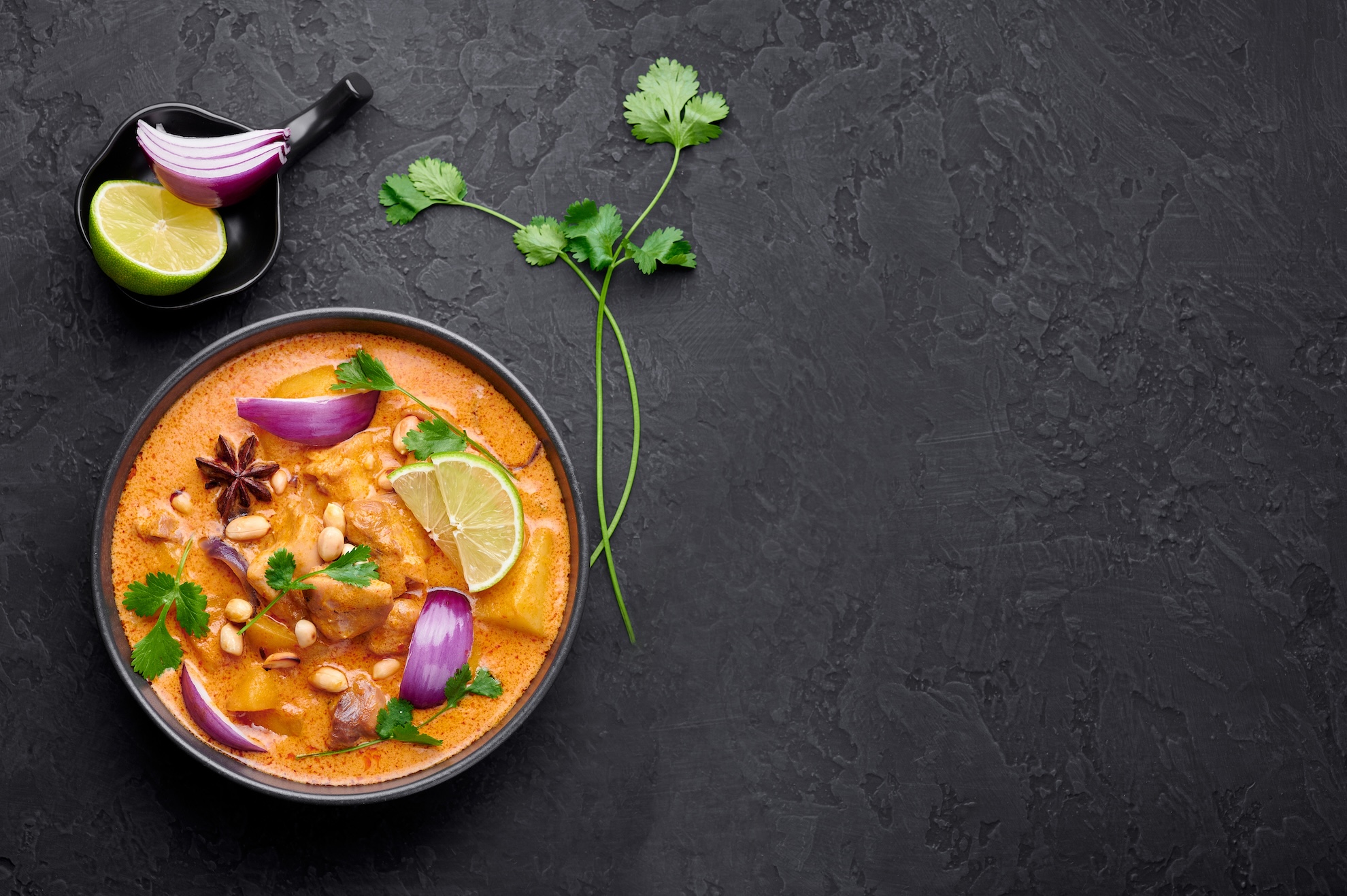 Thailändisches Curry in einer schwarzen Schale, mit Erdnüssen, Limetten, Zwiebeln und frischem Koriander. 